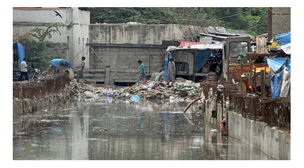 Flooded urban area with stagnant water, rubble, and debris. People walk on makeshift paths beside dilapidated structures and blue tarpaulin-covered stalls.