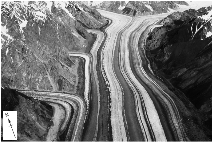 Aerial view of a winding glacier between snow-capped mountains, showing distinct layers of ice and rock, with a north arrow in the lower left corner.