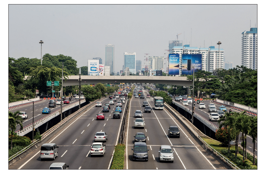 A busy multi-lane motorway lined with trees and high-rise buildings in the distance under a clear sky, showcasing urban traffic and billboards.