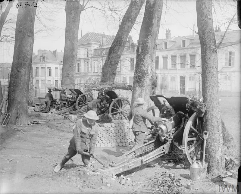 Soldiers operating artillery pieces in a wooded area with buildings in the background. Ammunition stacked nearby, with one soldier adjusting a cannon.