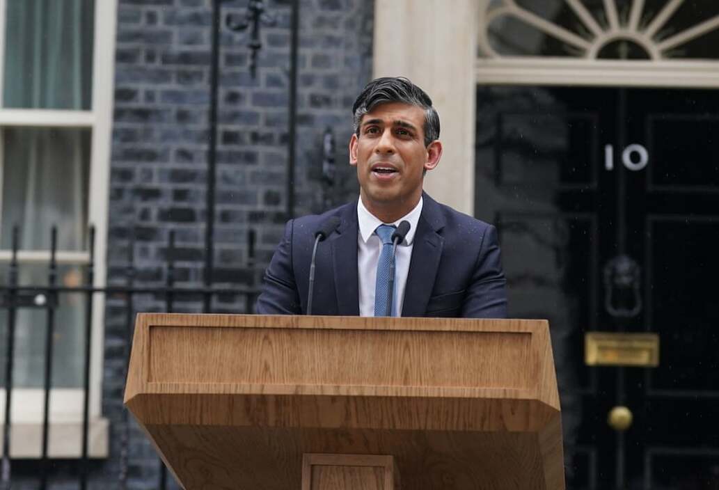 Man in a suit speaks at a wooden podium outside a building, with a black door and the number 10 visible in the background.