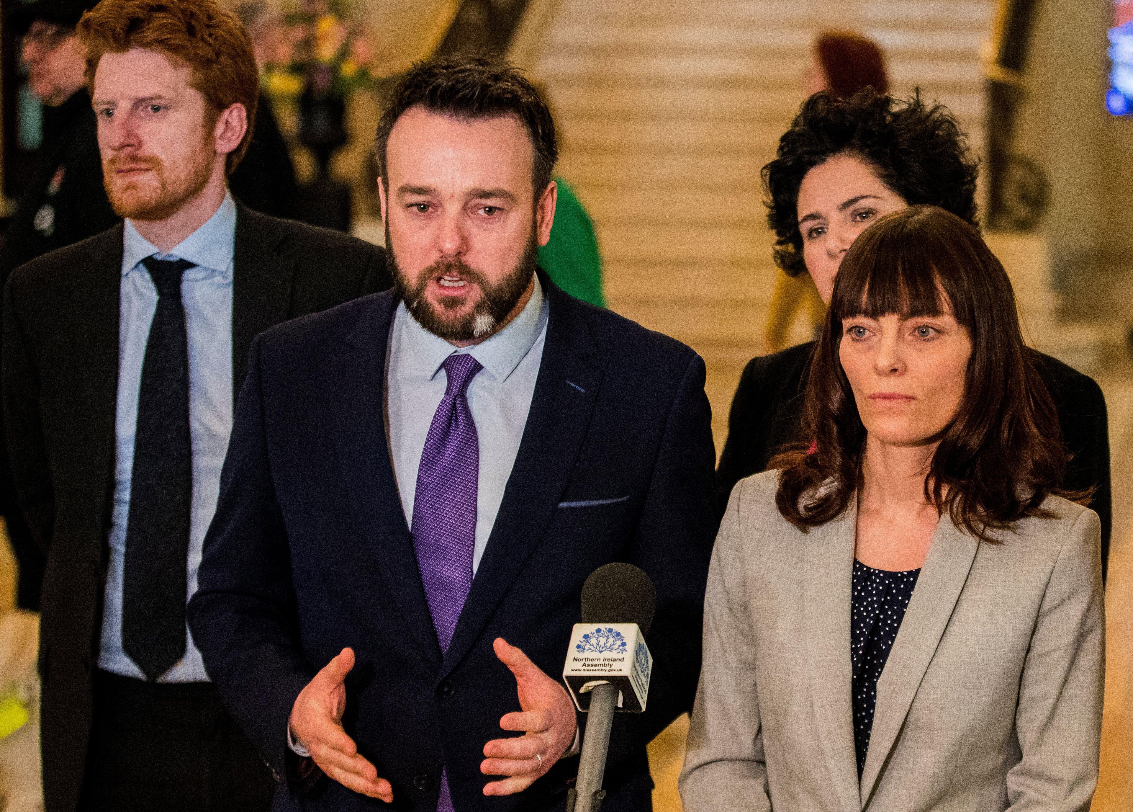 An MP stands before a media microphone, flanked by members of the public