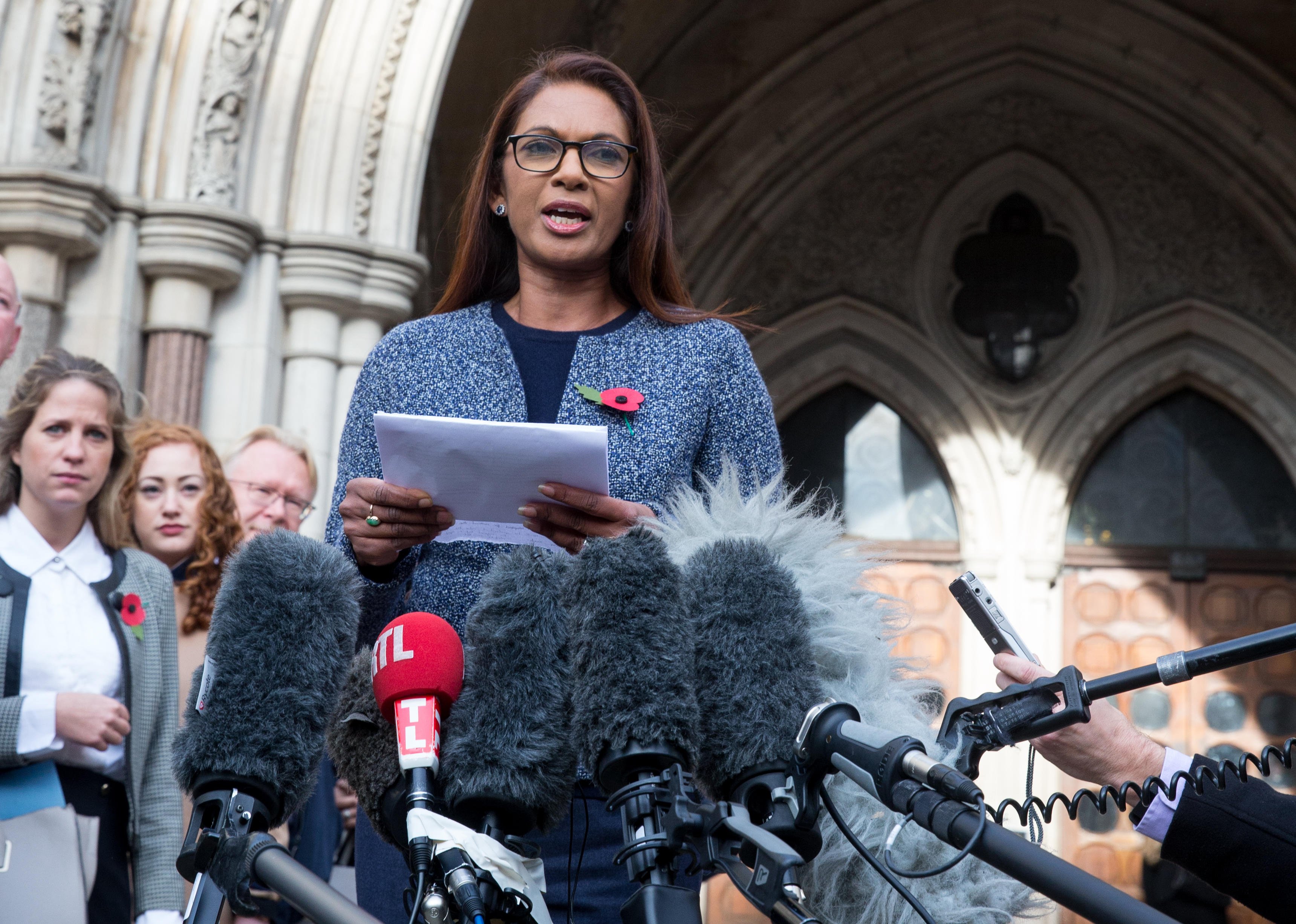 A woman speaks at a press conference outside a historic building, standing behind numerous microphones, with several people in the background.