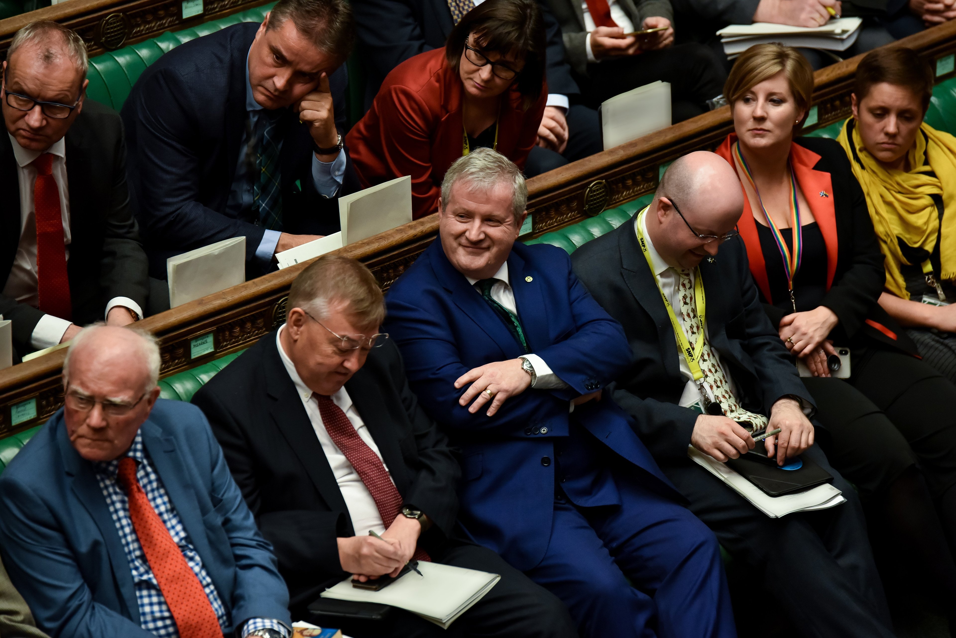 A smiling man in a blue suit sits on the green benches of the House of Commons, surrounded by Parliamentary colleagues