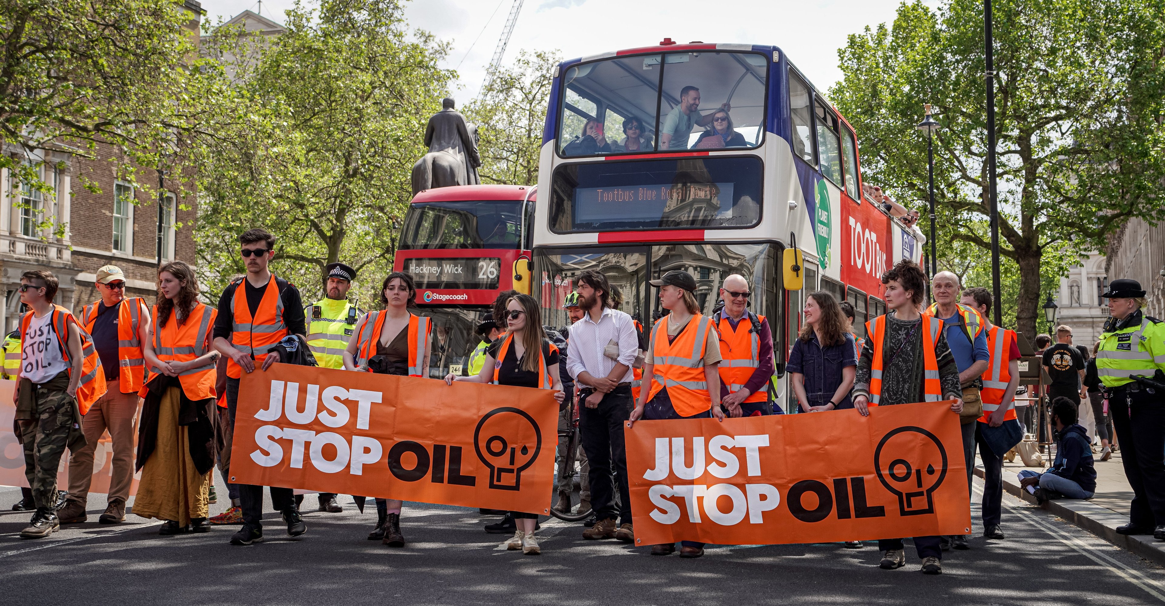 Protestors wearing hi-vis jackets with banners that say 'Just stop oil', standing in front of two buses on a tree-lined urban street