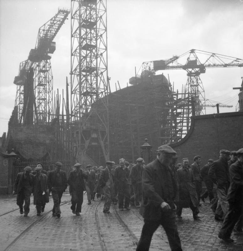 Workers leave a shipyard, with cranes and the hull of a ship under construction visible in the background, capturing an industrial scene.