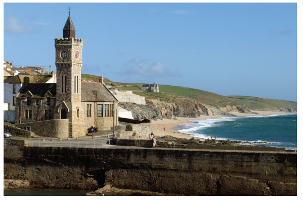 Historic stone church with a clock tower by a coastal cliff, overlooking a sandy beach and vibrant blue sea under a clear sky.