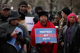 Protestors holding signs including "Protect My Vote" demonstrate outdoors in winter clothing, advocating for voting rights in a diverse group.