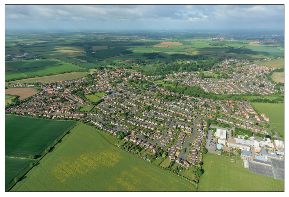 Aerial view of a small English village surrounded by green fields and farmland, showing winding streets with houses and a visible industrial area.