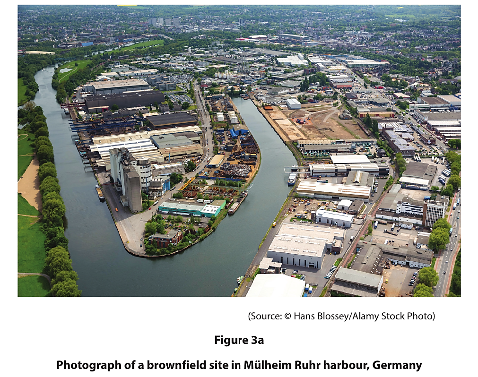 Aerial view of an industrial brownfield site by a river in Mülheim Ruhr harbour, Germany, featuring warehouses, factories, and greenery.