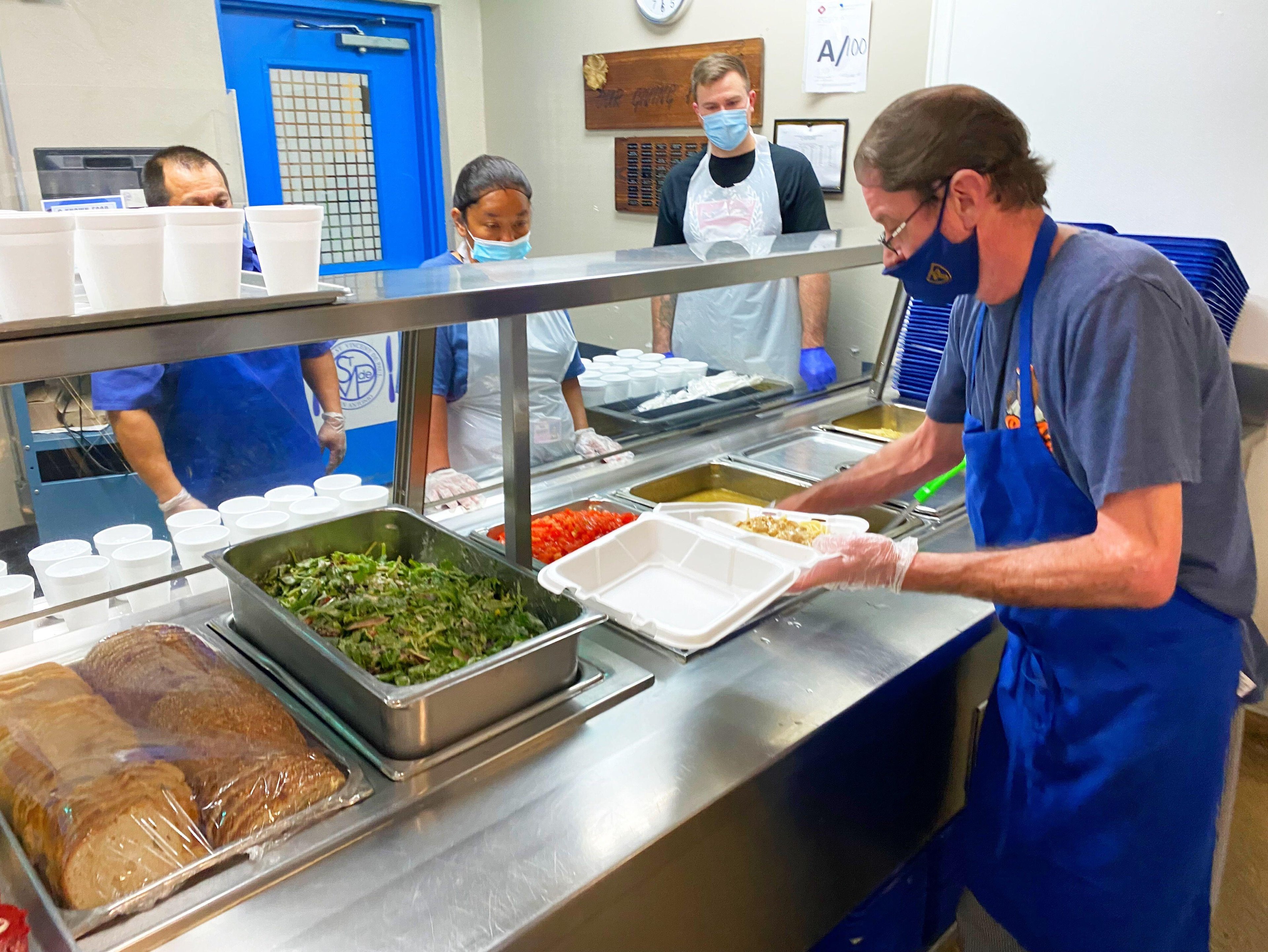 Volunteers in a kitchen serve food into takeaway containers, with trays of salad, bread, and other dishes visible under a glass counter.