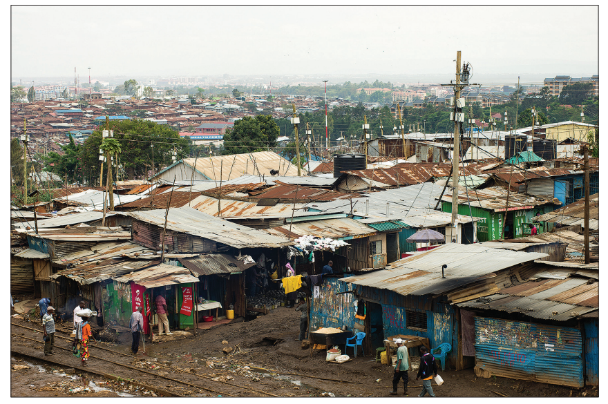 View of a densely populated informal settlement with tin-roofed shacks, scattered people, electrical poles, and urban skyline in the background.