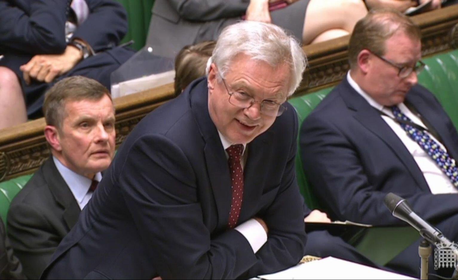 A man speaking at a microphone in a government assembly, surrounded by seated colleagues in suits, with papers on a desk in front of him.