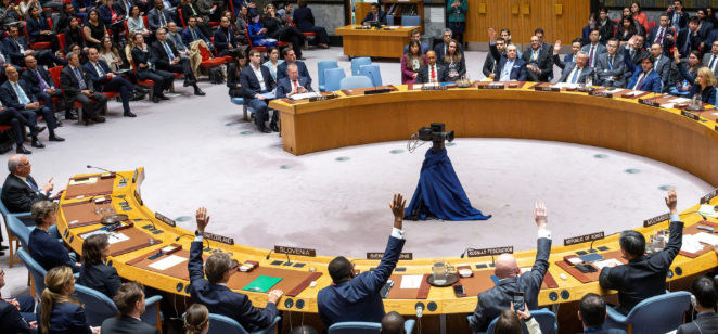 United Nations Security Council meeting, delegates in a circular seating arrangement, some raising hands to vote, with an overhead camera in the centre.