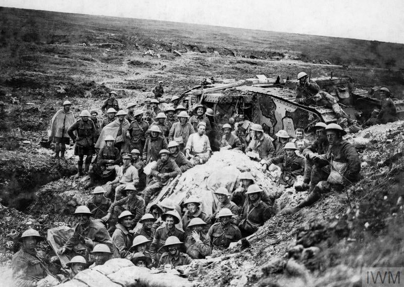 Soldiers in uniform rest in a trench beside a damaged tank on a battlefield, with barren land stretching behind them, during World War I.