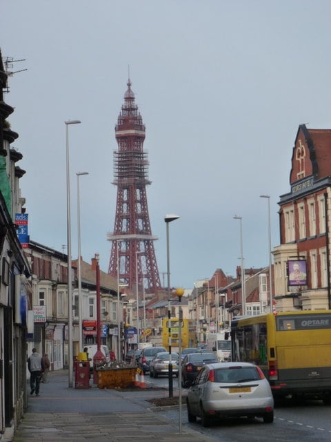 Street scene with vehicles, buildings, and pedestrians; a tall iron tower similar to the Eiffel Tower stands prominently in the background under a cloudy sky.