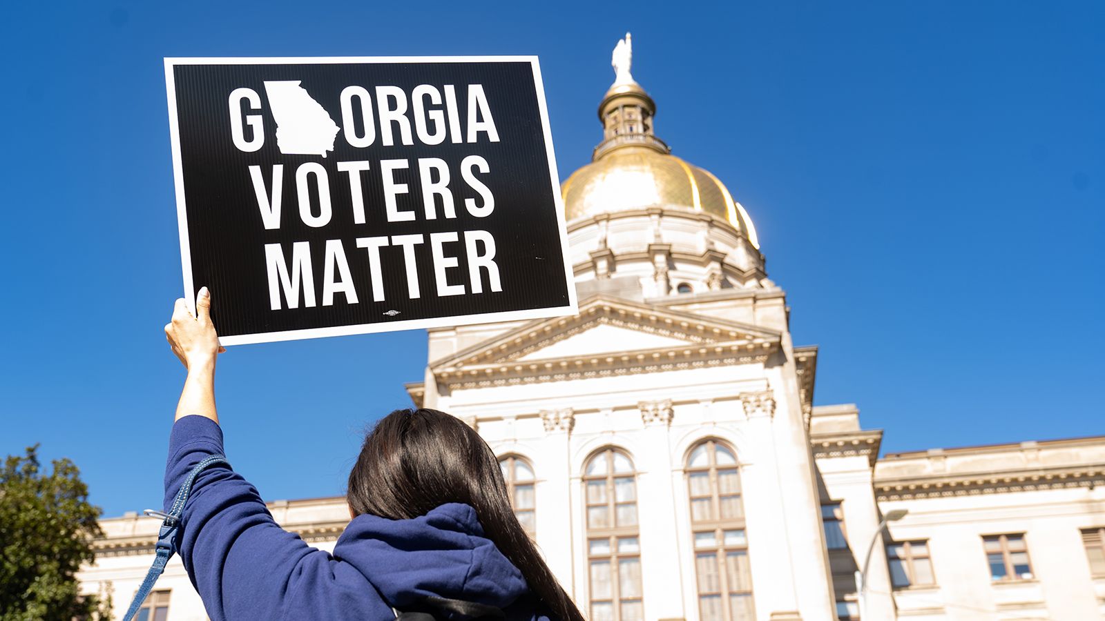 Person holds a sign reading "Georgia Voters Matter" in front of a historic building with a golden dome against a clear blue sky.