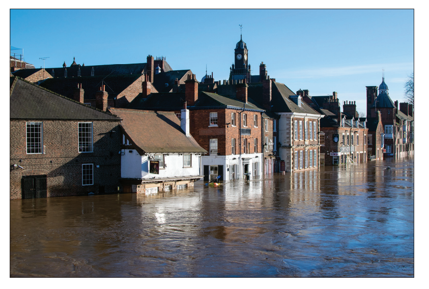 Flooded street in a town with row of brick buildings partially submerged, reflecting in the water under a clear blue sky. Clock tower visible in background.