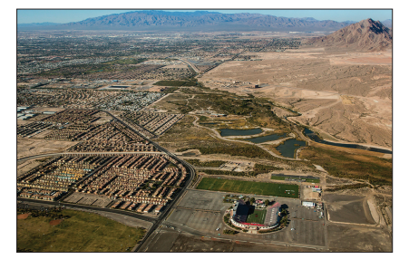 Aerial view of a desert cityscape with residential areas, roads, and a sports complex. In the distance, arid mountains under a clear blue sky.