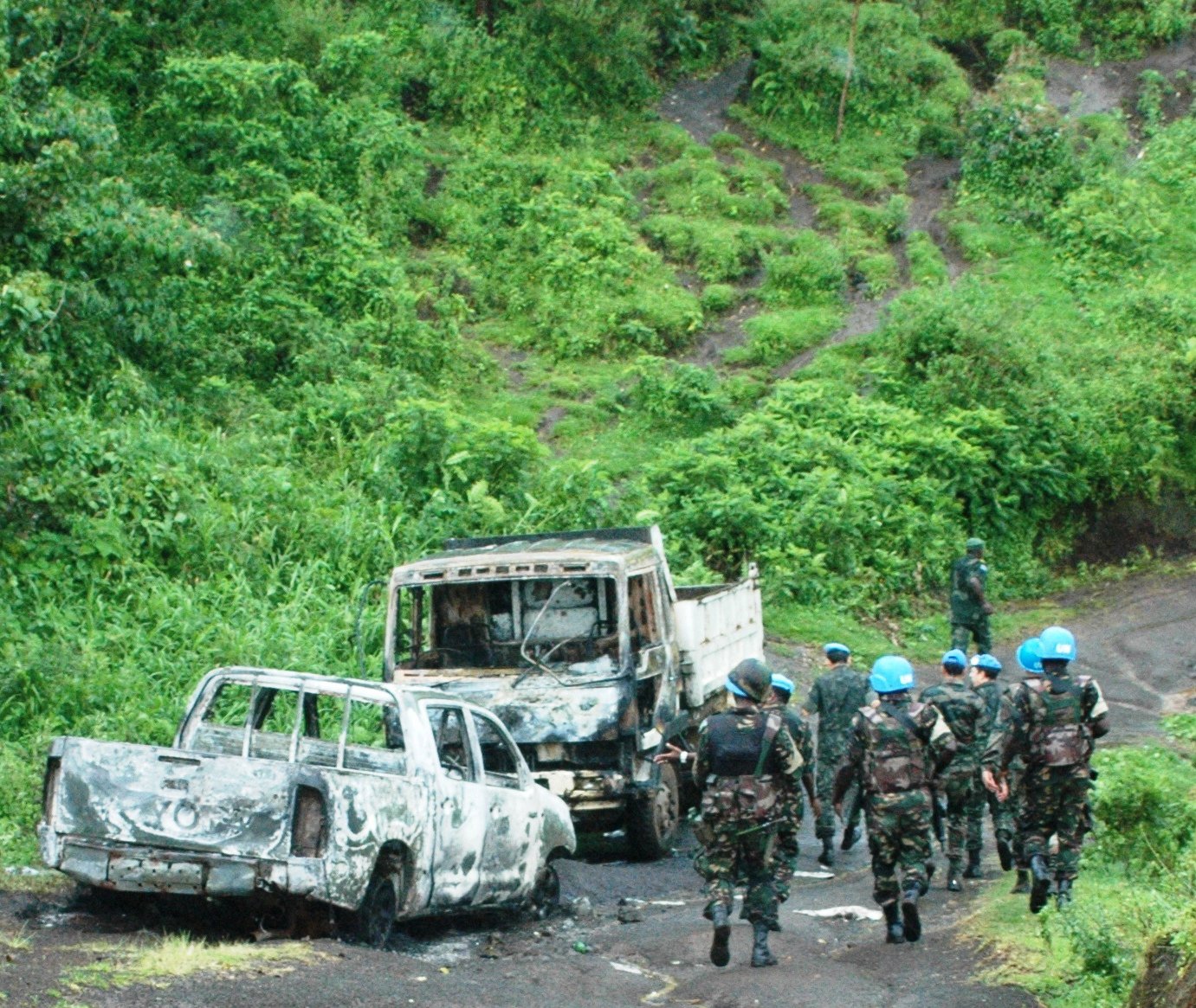 Soldiers in camouflage and blue helmets inspect burnt-out vehicles on a dirt road, surrounded by green hills and lush vegetation.