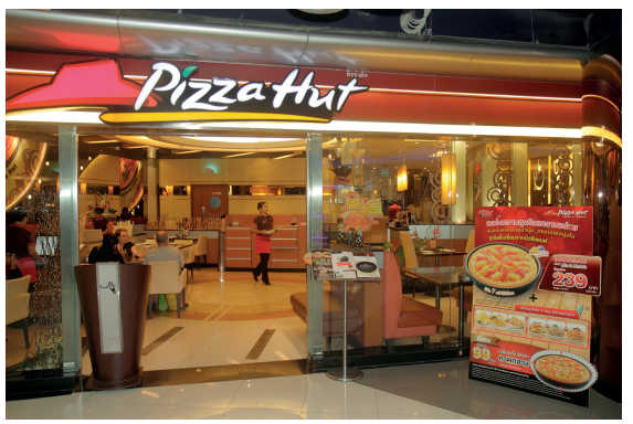 Front entrance of a Pizza Hut restaurant with illuminated signage, showcasing a promotional poster for pizza deals next to the glass doors.