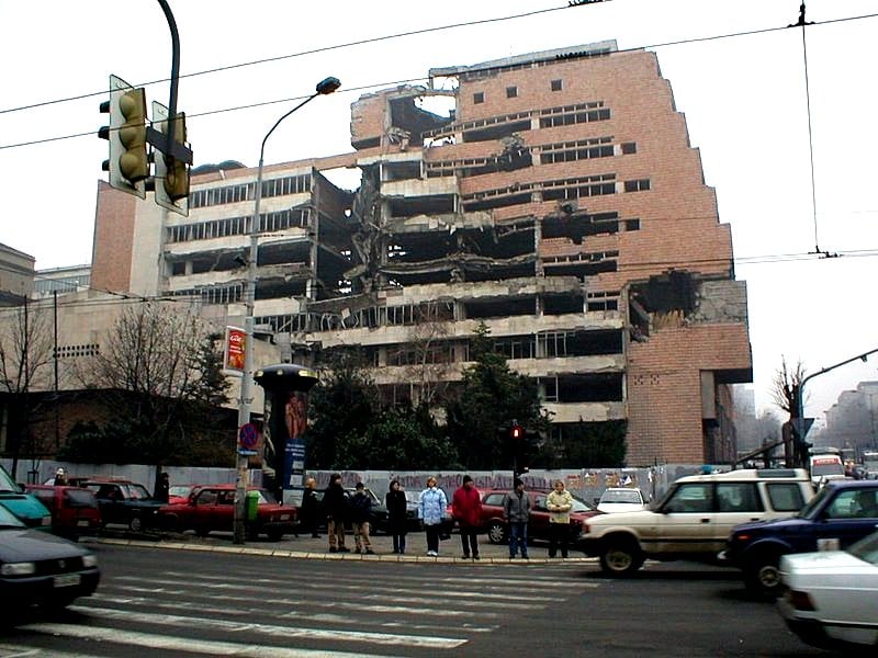 Damaged multi-storey building with exposed interior, crumbling walls, and debris. Cars and pedestrians cross a street in the foreground on a cloudy day.
