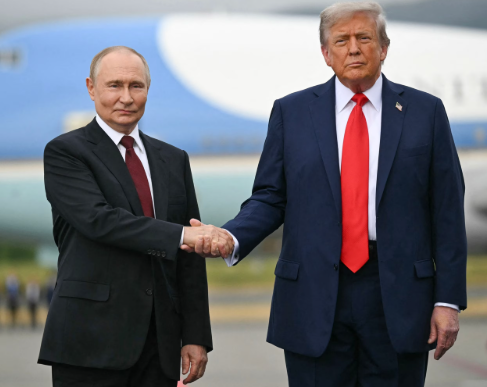 Two men in suits shake hands outside, with an aircraft partially visible in the background. One wears a red tie; the other wears a blue tie.