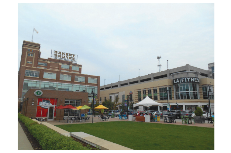 Outdoor plaza with green lawn, tables, and umbrellas in front of a brick building marked Bakery Square and a LA Fitness gym. Overcast sky.