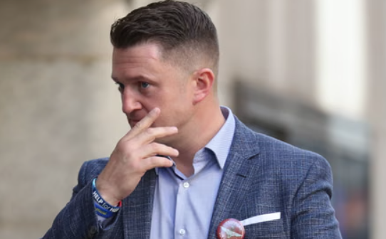 Man in blue blazer and light blue shirt touching his face, standing outdoors. Short hair, wearing wristbands and a button with a blurred background.