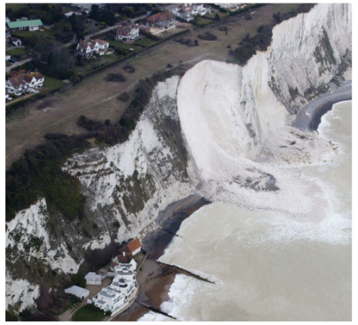 Aerial view of a coastal cliff collapse, showing white chalk cliffs, debris on the beach, and nearby houses with green fields in the background.