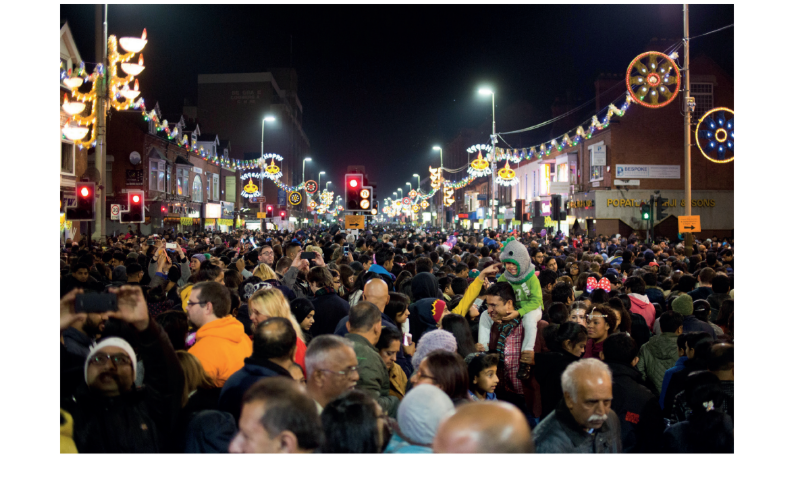 A bustling night street festival with a large crowd and vibrant hanging lights, people taking photos, and festive decorations above.