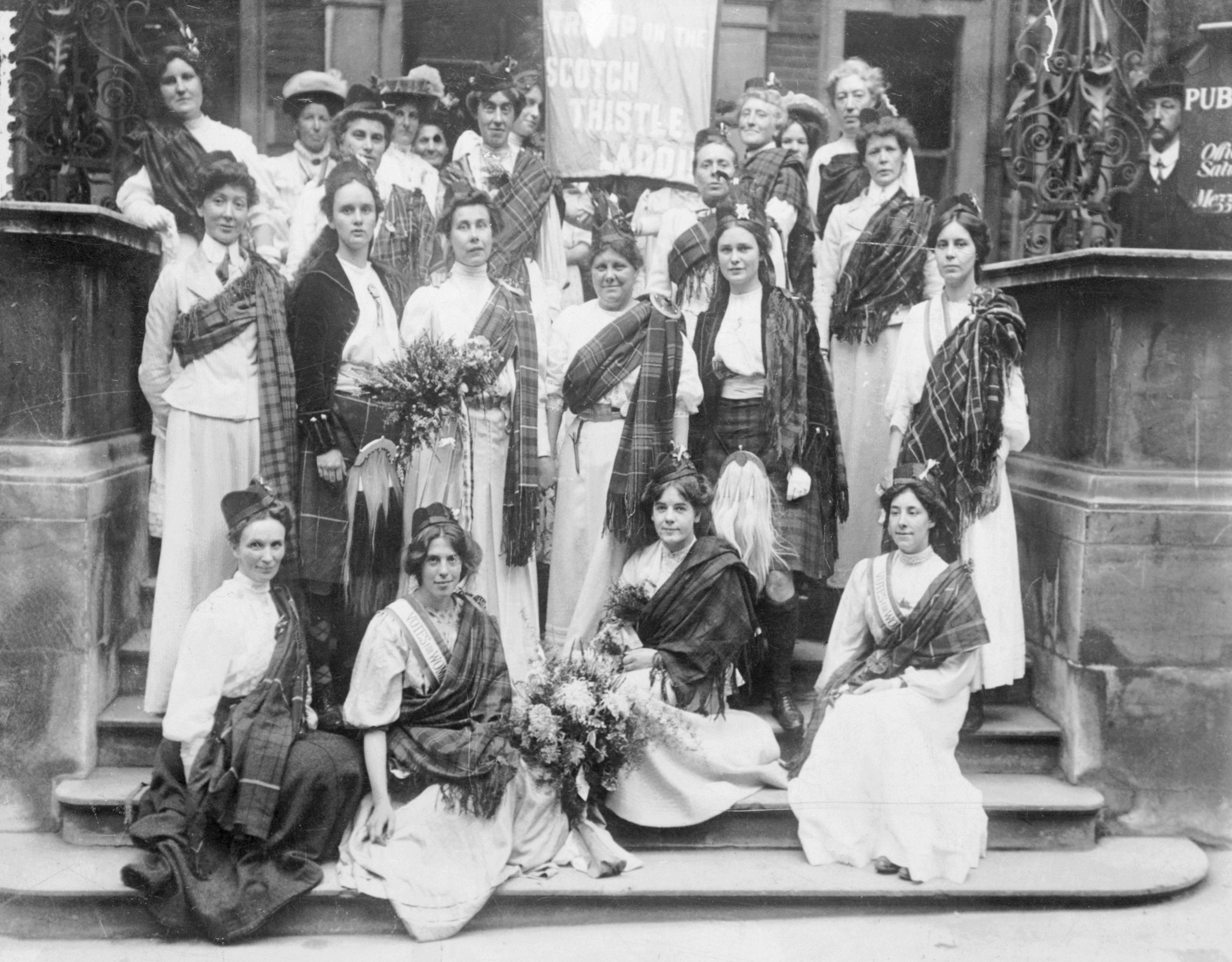 Group of women in period clothing, some wearing tartan shawls, posing on steps with bouquets. A banner and ornate pillars are visible in the background.