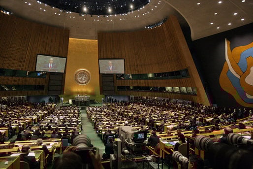 A large assembly hall with delegates seated, focusing on a speaker at a podium. Two screens display the speaker, with an emblem on a gold background behind.