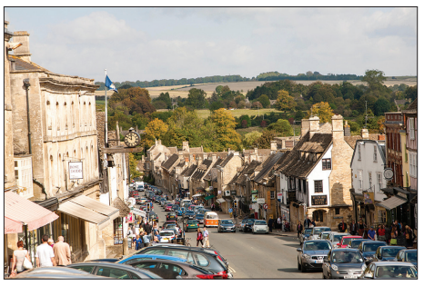 Busy street in a quaint village with historic stone buildings, cars parked along the road, and a scenic countryside backdrop under a partly cloudy sky.