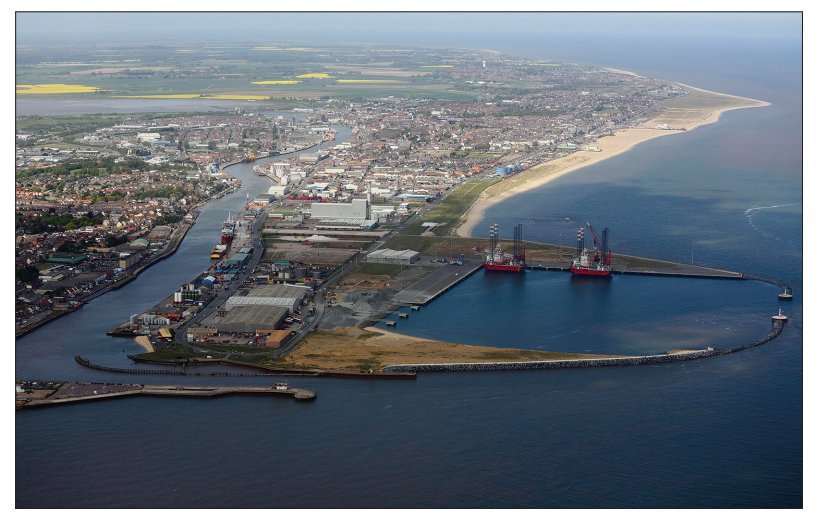 Aerial view of a coastal town with a prominent port area, featuring industrial buildings, cranes, two large ships, and a sandy beach lining the shoreline.