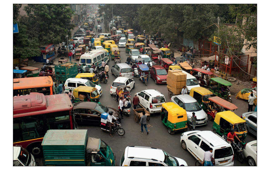 Busy city street with heavy traffic, including cars, buses, rickshaws, and motorbikes; pedestrians are weaving through vehicles, and trees line the road.