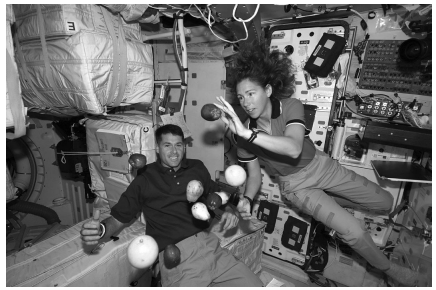 Two astronauts float in a space station module with several pieces of fruit suspended in zero gravity, surrounded by equipment and controls.