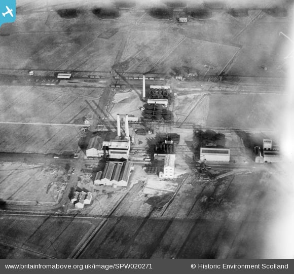 Aerial view of an industrial site with buildings, chimneys, and rail tracks amid farmlands, partially obscured by clouds.