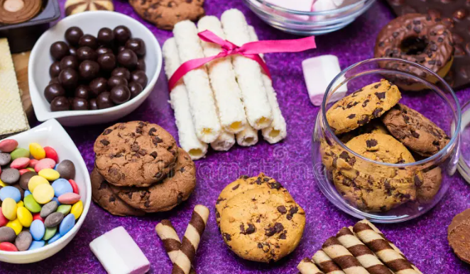 Assorted sweets on a purple background, including chocolate cookies, wafer rolls, candy, marshmallows, and chocolate balls in bowls and jars.