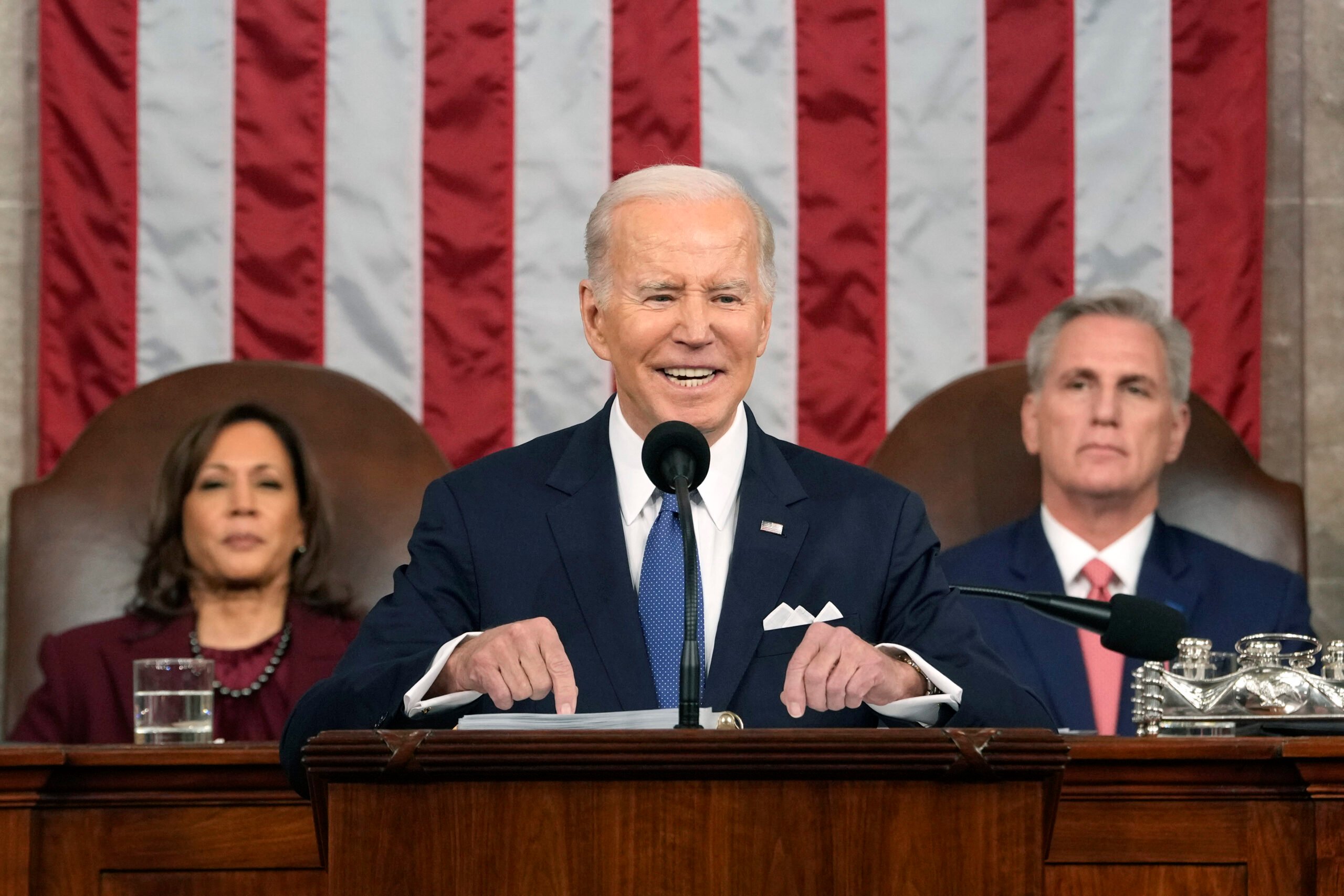 A man in a suit speaks at a podium with a microphone, flanked by two others sitting behind him, against a backdrop of a striped flag.