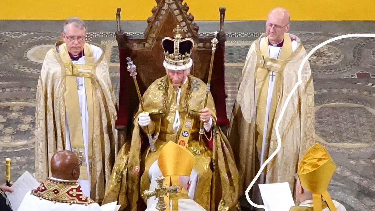 A man in ornate robes and crown sits on a throne holding sceptres, flanked by two clergymen in gold vestments, with ceremonial items on a table.