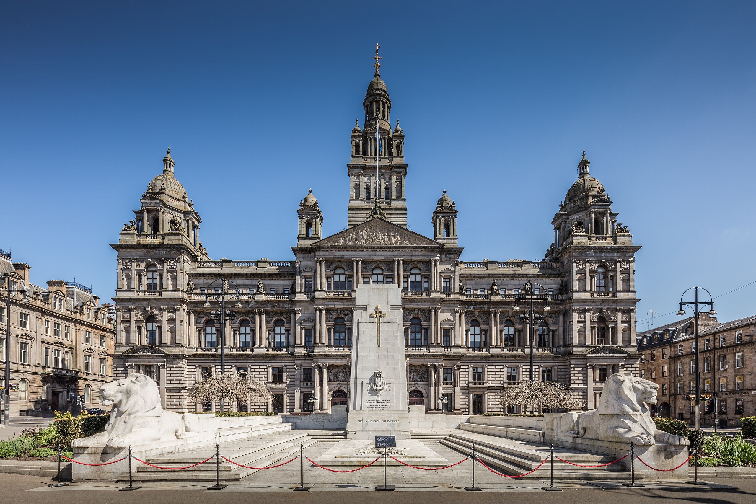 Historic building with ornate facade, central tower, flanking domes, and two large lion statues in front, set against a clear blue sky.