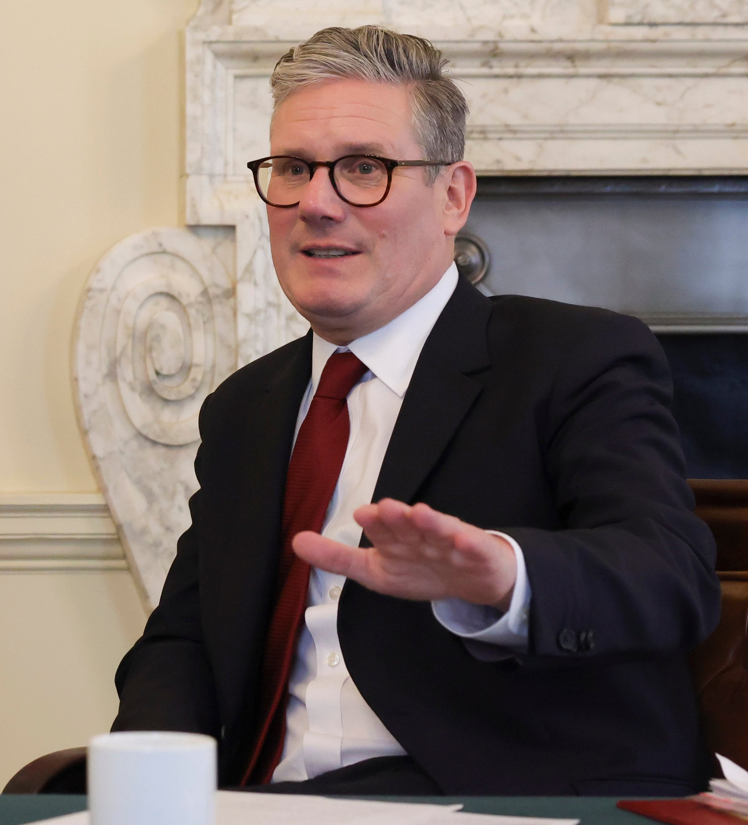 Man in a suit with glasses gesturing while sitting at a table, with a cup and papers in front of him, in a room with a marble fireplace.