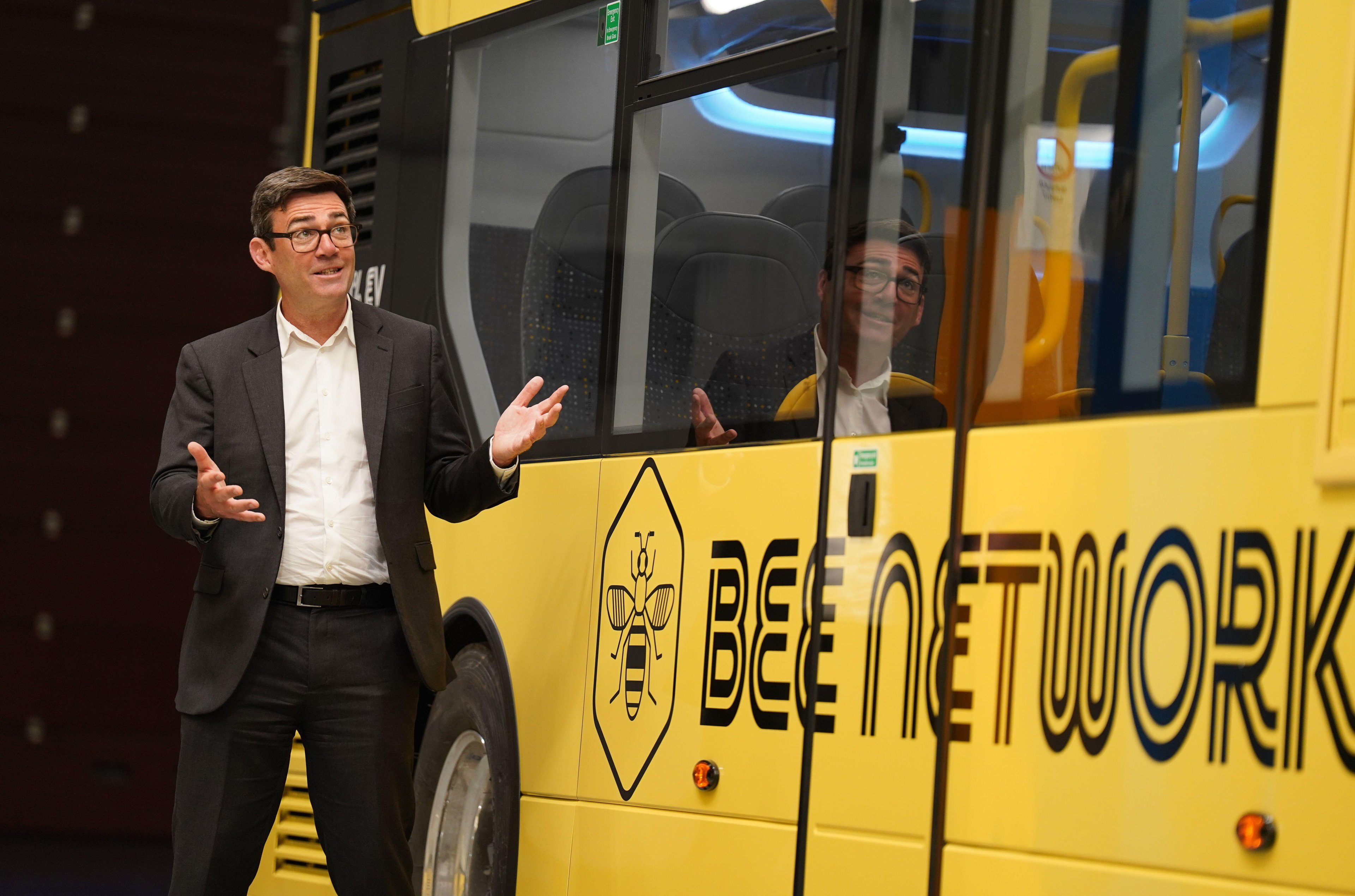 Man in a suit gestures next to a yellow bus with "Bee Network" and bee logo. Reflection visible in bus window. Interior seats and handles visible.