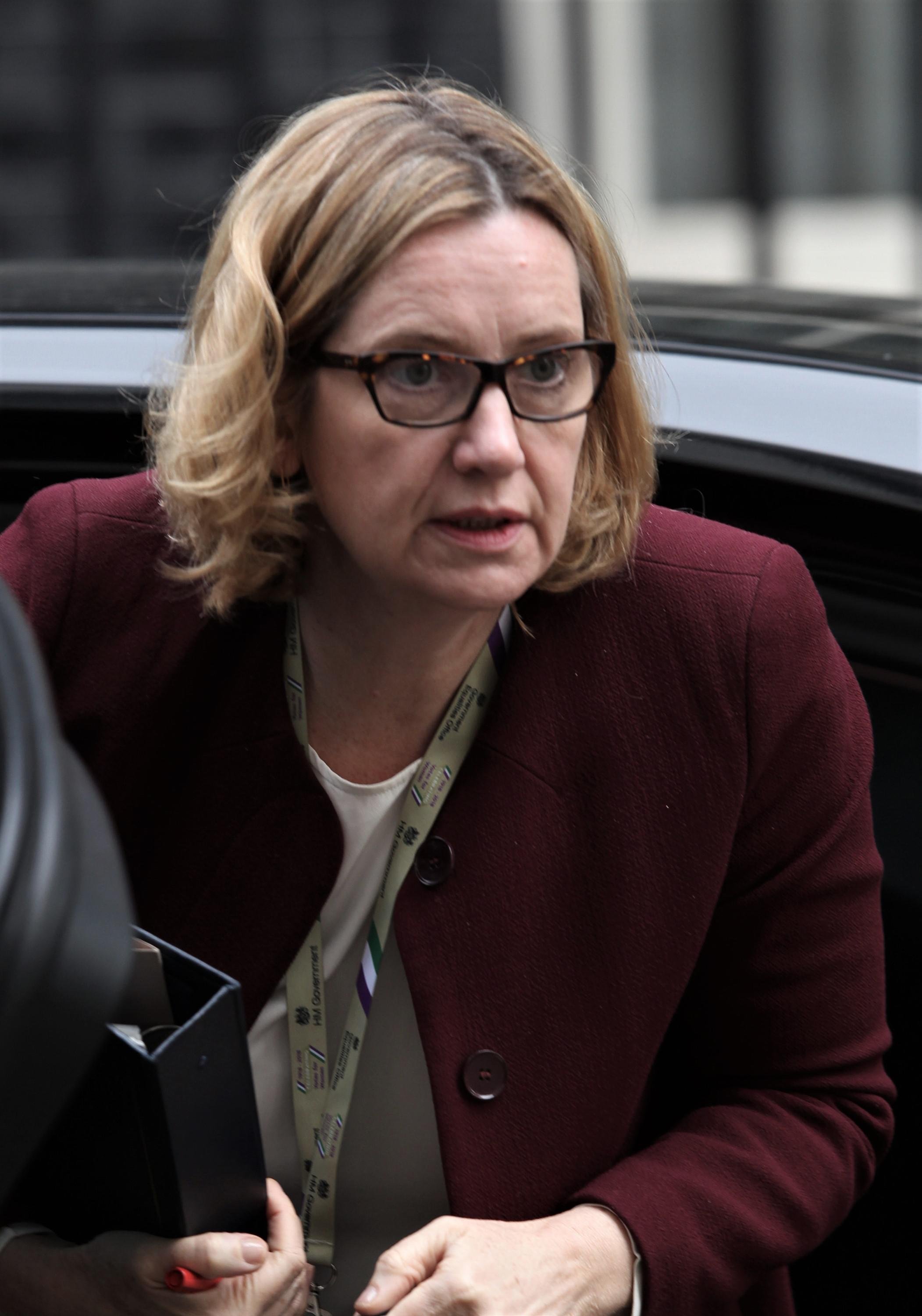 Woman with short blonde hair and glasses exiting a car, wearing a burgundy coat and a lanyard with ID badge, holding a folder and pen.
