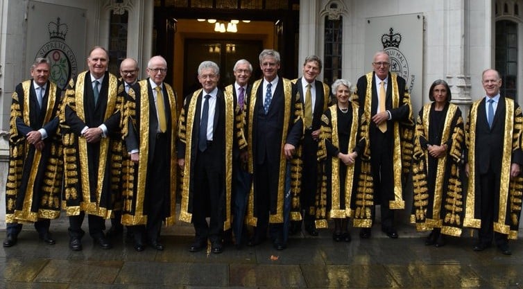 Group of judges in ceremonial robes stand in front of a grand building entrance, showcasing intricate gold detailing and official emblems.