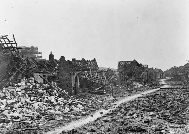 Rubble-lined street with severely damaged buildings, collapsed roofs, and debris scattered across the ground, creating a desolate war-torn scene.