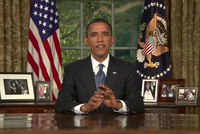 Man in a suit sits at a desk with family photos, flanked by American flags and a presidential seal, speaking with a serious expression.