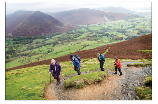 Four people hiking up a grassy hill with trekking poles, overlooking a scenic valley and distant hills under a cloudy sky.
