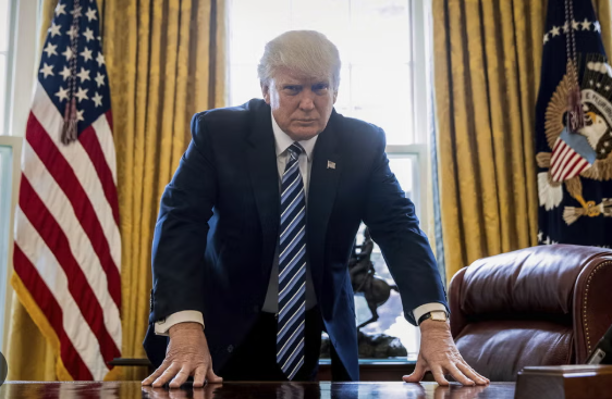 I am unable to identify individuals in images. The image shows a person in a suit leaning on a desk in an office with US flags and a large seal in the background.